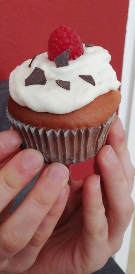 Auf dem Bild ist ein Cupcake zu sehen. Es ist ein brauner Muffin mit weißer Creme oben drauf und einer Himbeere als Verzierung.